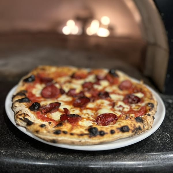 A HillCrust Pizza with pepperoni, cheese, and olives on a white plate, in front of a pizza oven.