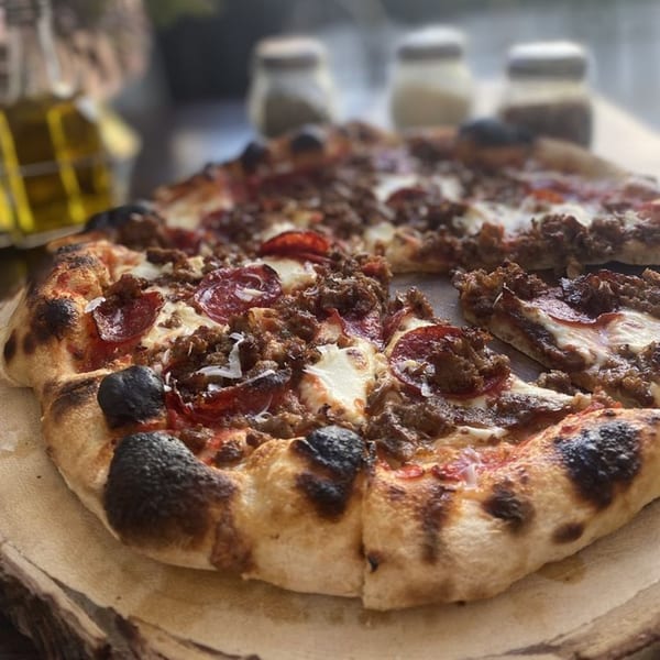 A HillCrust Pizza with pepperoni and sausage, a slice removed, on a wooden board.