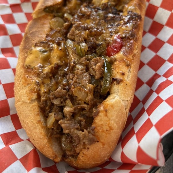 A HillCrust Pizza cheesesteak sandwich, overflowing with meat, onions, peppers, and melted cheese on a red checkered liner.