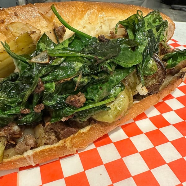A close-up of a HillCrust Pizza cheesesteak with spinach, onions, and melted cheese on a red and white checkered paper.